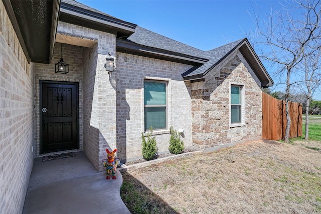 entrance to property featuring brick siding, roof with shingles, and fence