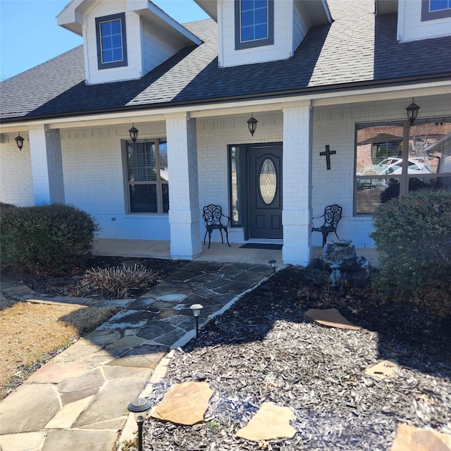 doorway to property with brick siding, covered porch, and roof with shingles