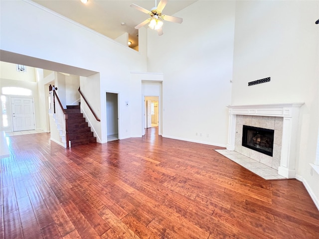 unfurnished living room featuring baseboards, ceiling fan, a tiled fireplace, stairway, and wood finished floors