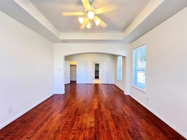 spare room with a raised ceiling, wood finished floors, arched walkways, baseboards, and ceiling fan