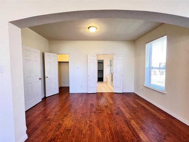 unfurnished bedroom featuring a spacious closet, dark wood-type flooring, baseboards, ensuite bathroom, and arched walkways