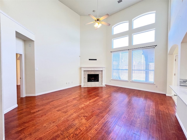 unfurnished living room with baseboards, a tiled fireplace, hardwood / wood-style floors, a towering ceiling, and a ceiling fan