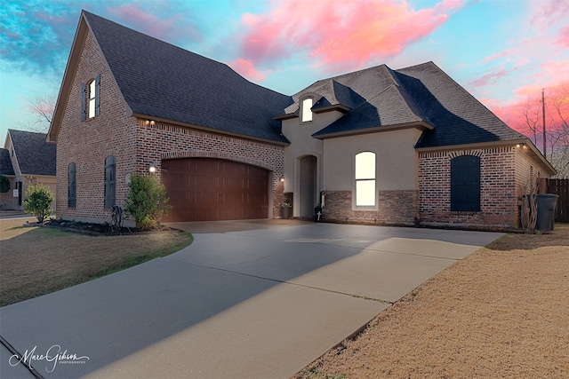 french country inspired facade with stucco siding, driveway, a shingled roof, a garage, and brick siding