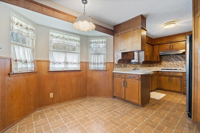 kitchen featuring under cabinet range hood, brown cabinetry, wainscoting, and light countertops