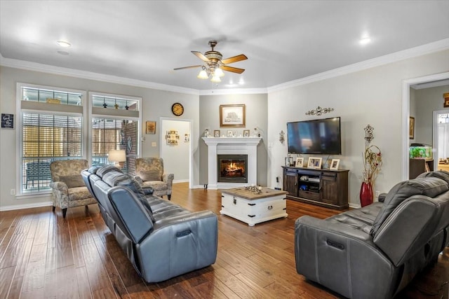 living room with dark wood-type flooring, ceiling fan, baseboards, ornamental molding, and a warm lit fireplace