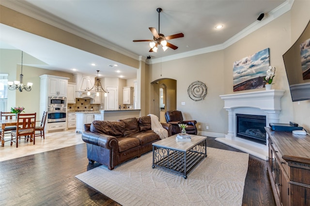 living room with wood finished floors, arched walkways, a glass covered fireplace, crown molding, and ceiling fan with notable chandelier