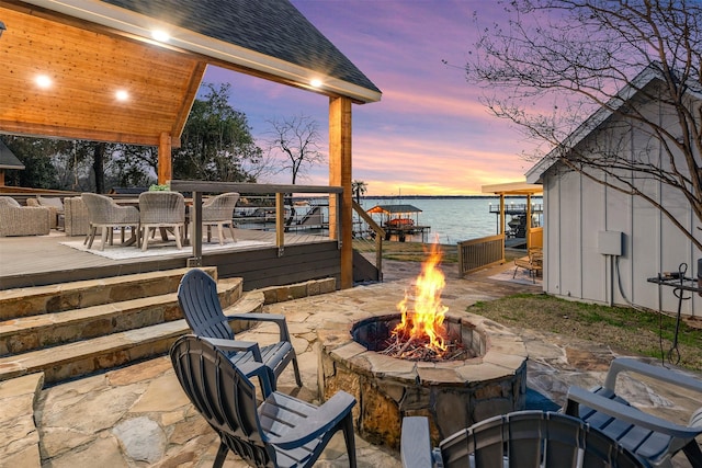 patio terrace at dusk with outdoor dining area, a deck with water view, and an outdoor fire pit