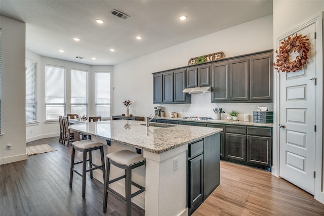 kitchen with under cabinet range hood, stainless steel gas cooktop, light wood-style flooring, a kitchen breakfast bar, and a sink