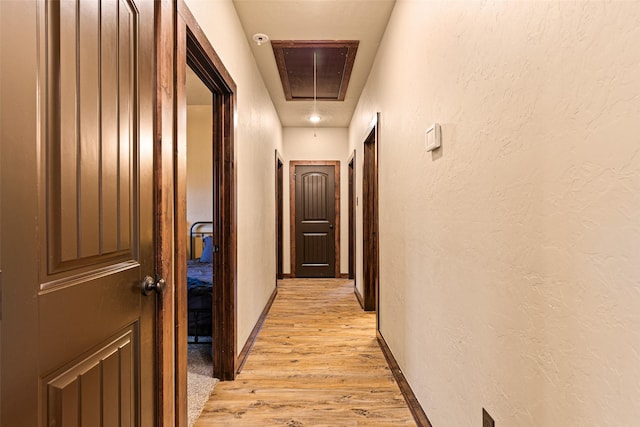 corridor featuring attic access, light wood-type flooring, baseboards, and a textured wall