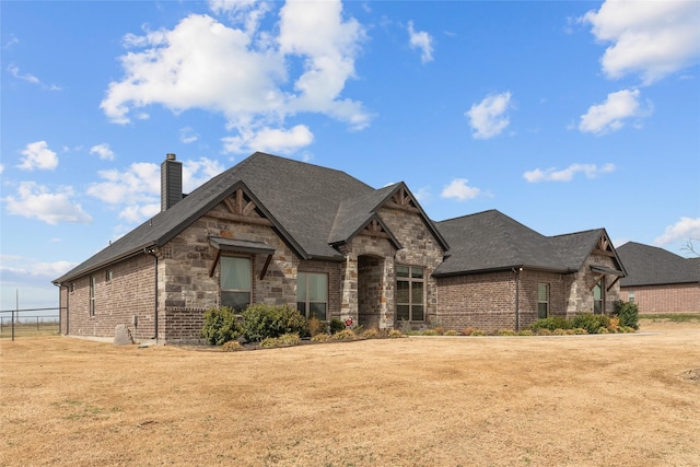 french country inspired facade featuring a front lawn, stone siding, a shingled roof, brick siding, and a chimney