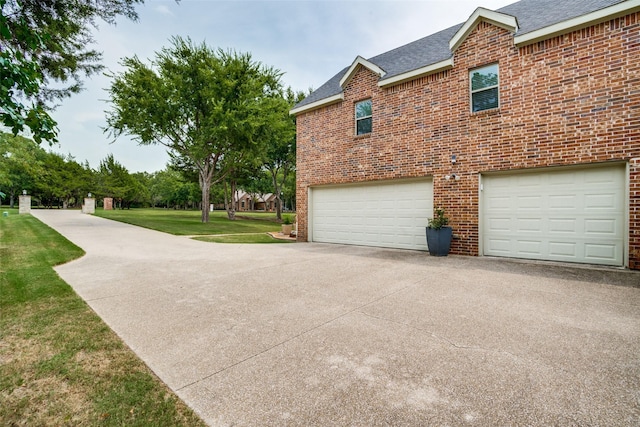 view of side of home featuring driveway, roof with shingles, a garage, a lawn, and brick siding