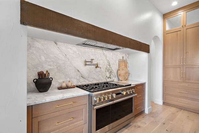 kitchen with light stone countertops, light wood finished floors, stainless steel stove, glass insert cabinets, and tasteful backsplash