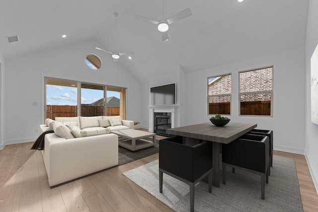 living area with visible vents, high vaulted ceiling, a glass covered fireplace, light wood finished floors, and ceiling fan