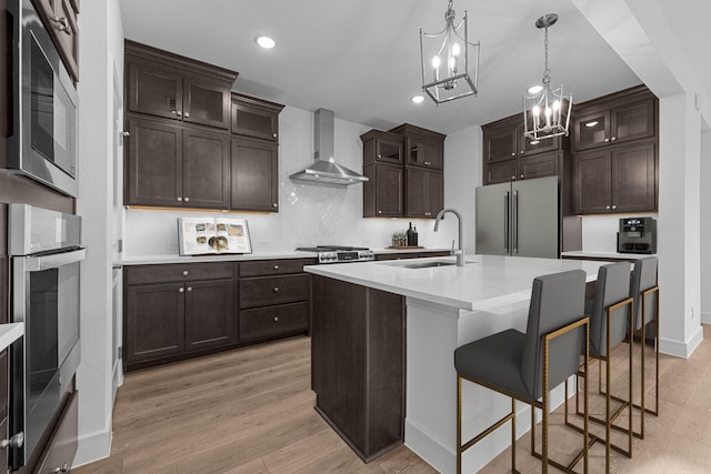 kitchen featuring light wood finished floors, wall chimney range hood, light countertops, appliances with stainless steel finishes, and a sink