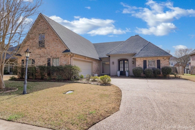 french country inspired facade with brick siding, a front yard, roof with shingles, driveway, and an attached garage