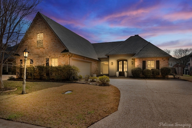 french country home with an attached garage, brick siding, driveway, and roof with shingles