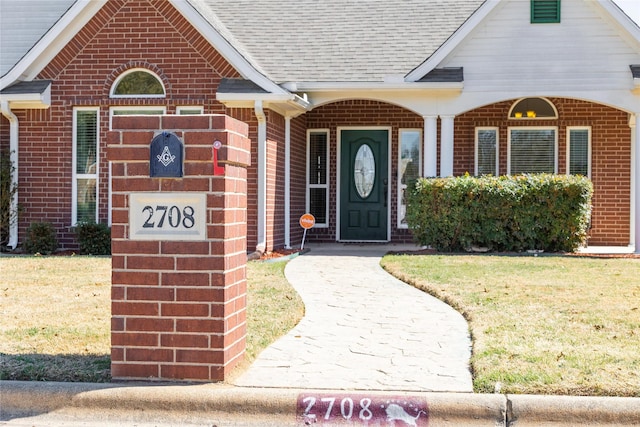 entrance to property with a yard, brick siding, and roof with shingles