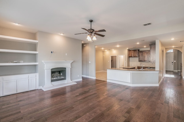 unfurnished living room featuring a ceiling fan, arched walkways, dark wood-style flooring, a sink, and a fireplace with raised hearth