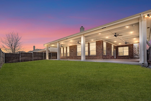 back of house at dusk with brick siding, a fenced backyard, a ceiling fan, and a lawn