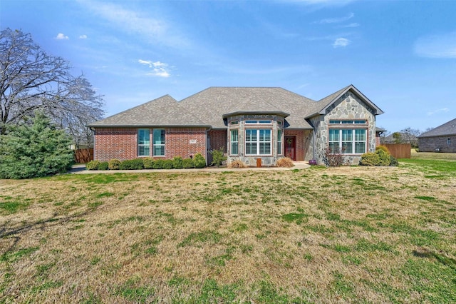 view of front facade with brick siding, a front lawn, fence, roof with shingles, and stone siding