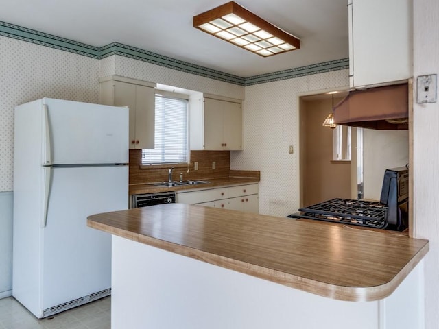 kitchen featuring a sink, freestanding refrigerator, a peninsula, wallpapered walls, and light floors