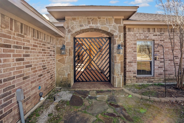 view of exterior entry featuring a gate, brick siding, stone siding, and a shingled roof