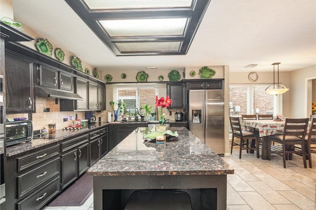 kitchen featuring stainless steel refrigerator with ice dispenser, under cabinet range hood, backsplash, black electric cooktop, and dark cabinets