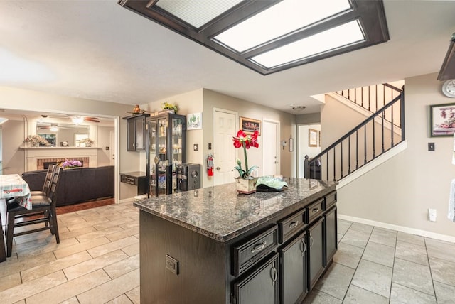 kitchen with dark stone countertops, a center island, a fireplace, baseboards, and ceiling fan