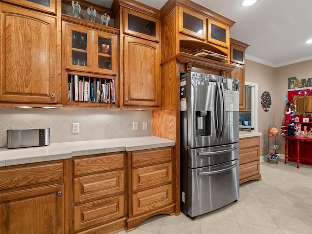 kitchen featuring stainless steel fridge with ice dispenser, glass insert cabinets, crown molding, brown cabinets, and backsplash