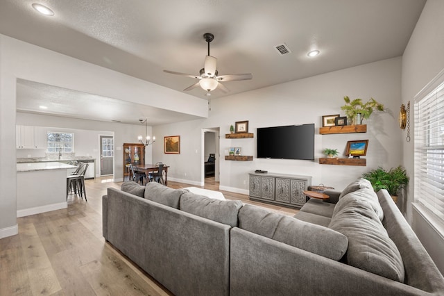living area featuring baseboards, a wealth of natural light, light wood-style floors, visible vents, and ceiling fan with notable chandelier