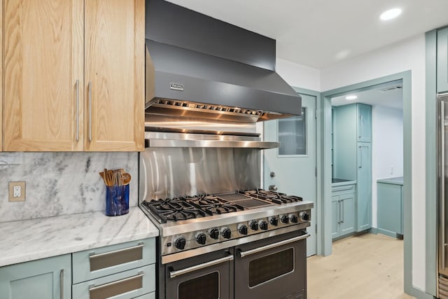 kitchen featuring light stone countertops, range with two ovens, wall chimney exhaust hood, light wood-type flooring, and backsplash