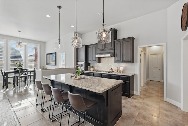 kitchen featuring an island with sink, a sink, decorative backsplash, stainless steel gas stovetop, and under cabinet range hood