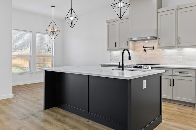 kitchen with light stone countertops, light wood-style flooring, a kitchen island with sink, decorative light fixtures, and backsplash