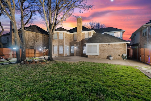 back of property at dusk featuring brick siding, a lawn, a patio, and fence