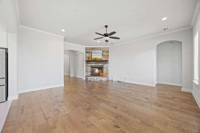 unfurnished living room featuring visible vents, light wood finished floors, arched walkways, a stone fireplace, and crown molding