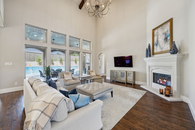 living area with baseboards, an inviting chandelier, a high ceiling, dark wood-style flooring, and a glass covered fireplace