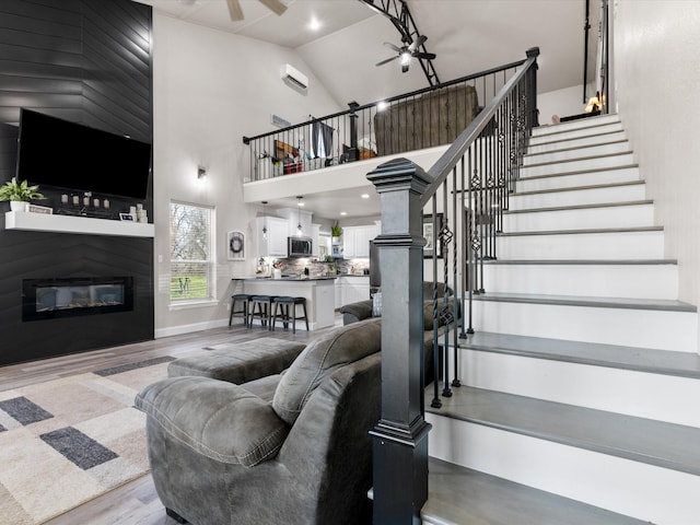 living area featuring stairs, wood finished floors, a glass covered fireplace, high vaulted ceiling, and a ceiling fan