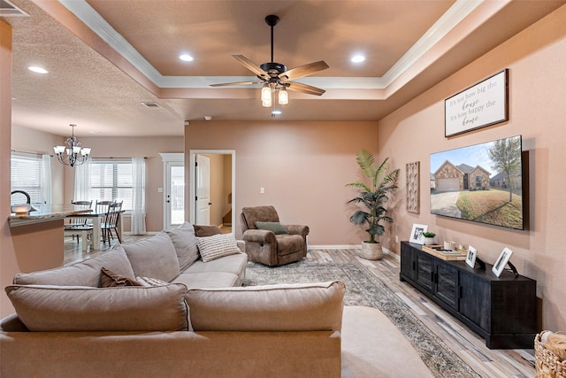 living area with ceiling fan with notable chandelier, a tray ceiling, crown molding, and light wood finished floors