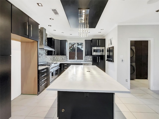 kitchen with light countertops, wall chimney exhaust hood, dark cabinetry, stainless steel appliances, and stacked washer / drying machine