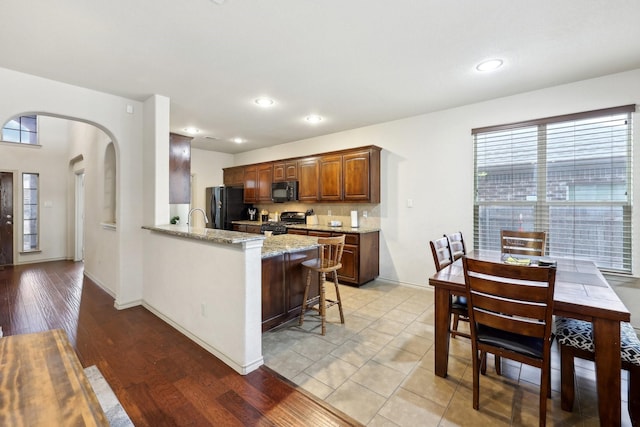 kitchen with black appliances, light stone counters, a kitchen breakfast bar, arched walkways, and a peninsula