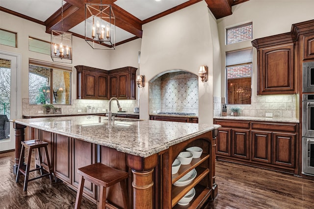 kitchen featuring light stone countertops, beam ceiling, a kitchen island with sink, dark wood-style flooring, and stainless steel appliances