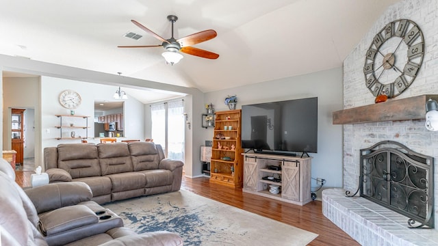 living area featuring a ceiling fan, wood finished floors, visible vents, vaulted ceiling, and a brick fireplace