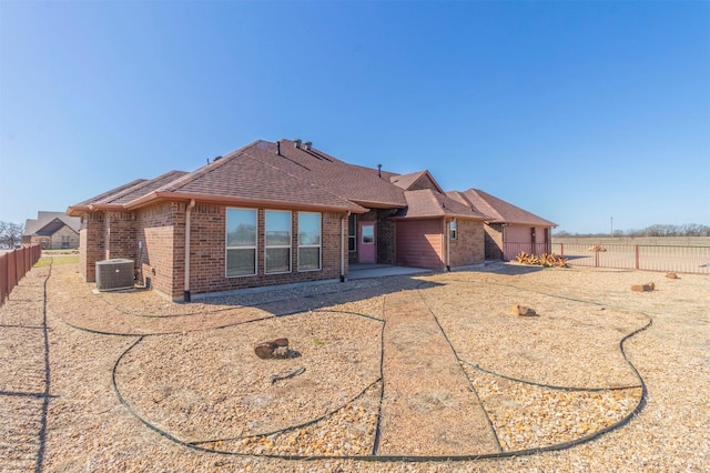 back of property with central AC unit, fence, brick siding, and roof with shingles