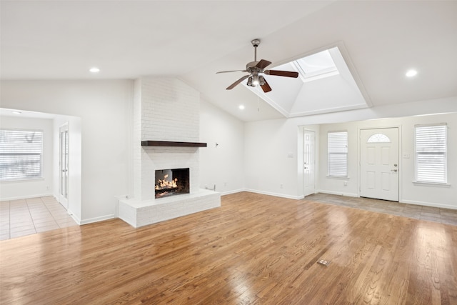 unfurnished living room with baseboards, ceiling fan, light wood-type flooring, vaulted ceiling with skylight, and a fireplace