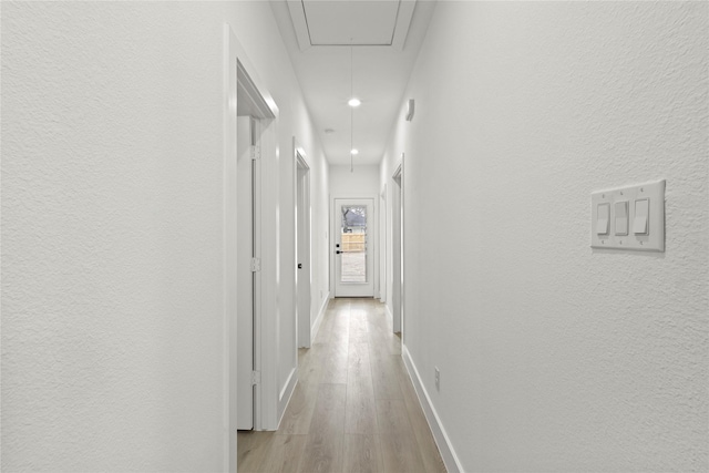 corridor with baseboards, attic access, light wood-style flooring, and a textured wall