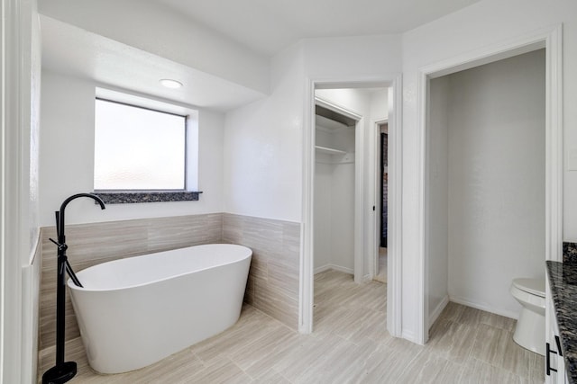 bathroom featuring a spacious closet, toilet, a freestanding bath, and tile walls