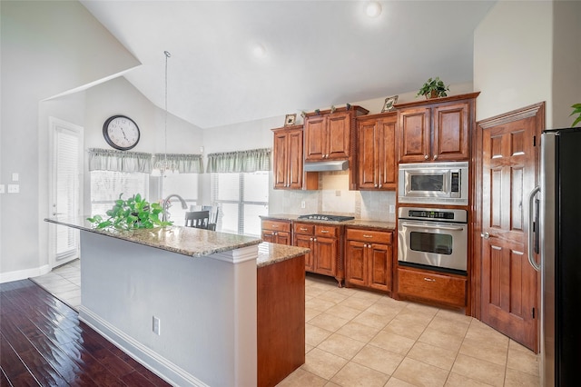 kitchen with light stone countertops, under cabinet range hood, brown cabinets, stainless steel appliances, and a kitchen island with sink