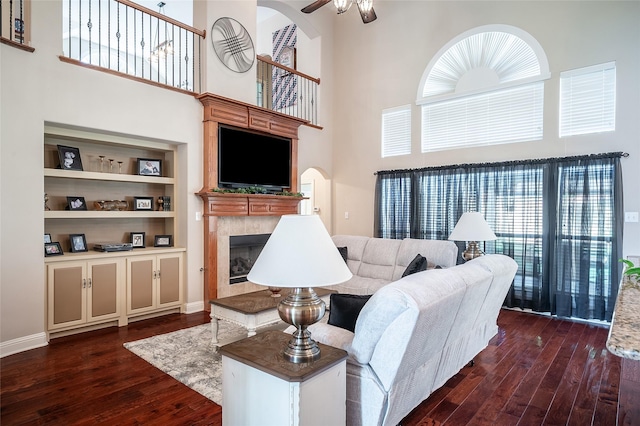 living area with built in shelves, a fireplace, dark wood-style flooring, ceiling fan, and a towering ceiling