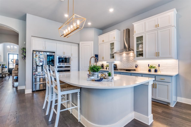 kitchen with an island with sink, arched walkways, stainless steel appliances, decorative backsplash, and wall chimney range hood