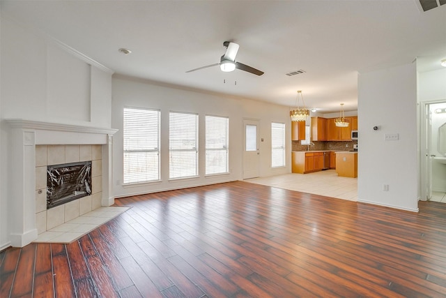 unfurnished living room featuring visible vents, a tiled fireplace, ornamental molding, light wood-style flooring, and ceiling fan with notable chandelier